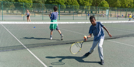 Youth playing tennis at Finsbury Park. Boy in foreground is about to hit a tennis ball with his racquet.