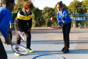 Girl bounces a tennis ball on a tennis racket while another girl and their coach watch